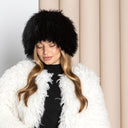 A woman wearing a white fur coat and a black furry hat stands in front of a beige curtain.