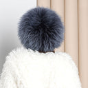 A close-up view of a person wearing a white fur coat with a fluffy gray pom pom on top.