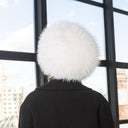 A person wearing a black coat and a white furry hat is standing in front of a window, looking out at a cityscape.